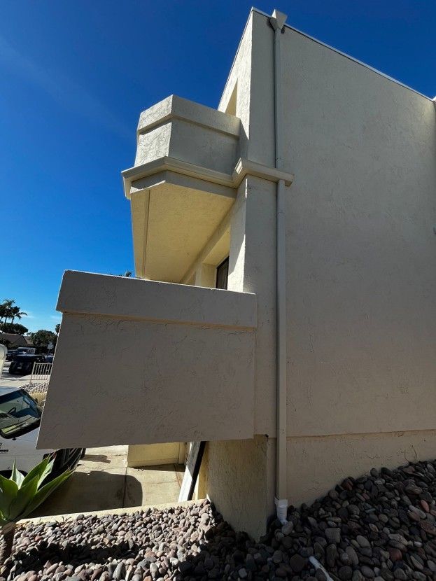 Beige building corner with a protruding balcony and gravel landscaping under a clear blue sky