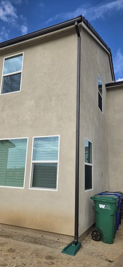 Side of a beige house with windows, a green trash bin, and a broom by the wall.