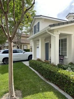 Suburban house exterior with white trim, a silver car in the driveway, and a green lawn lined by shrubs.