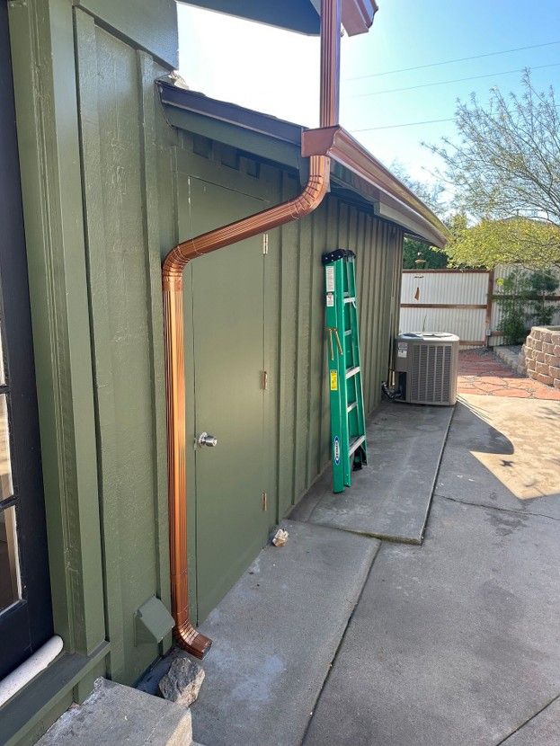 Green building side with a copper downspout, ladder, and HVAC unit along a concrete walkway.