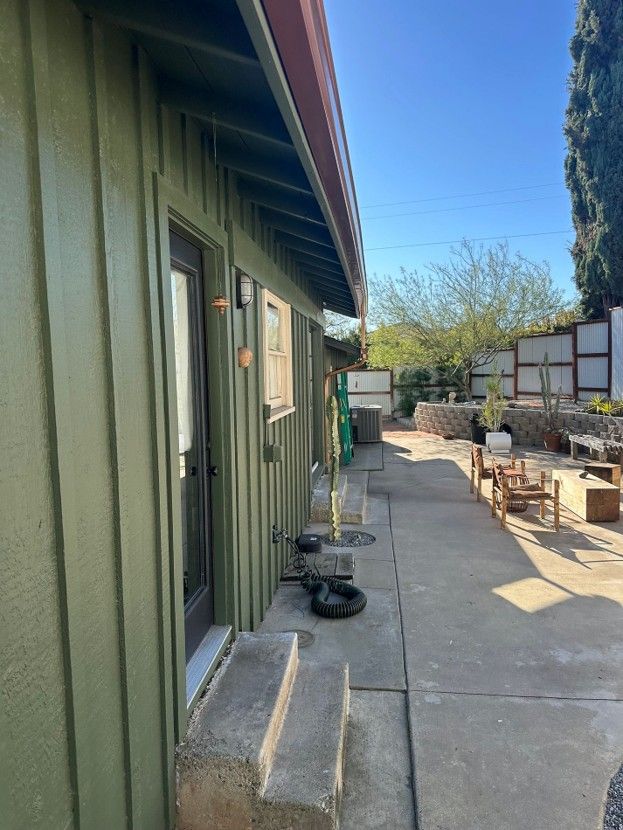 Side yard walkway beside a green house, with steps, a door, potted plants, and a sunny patio area.