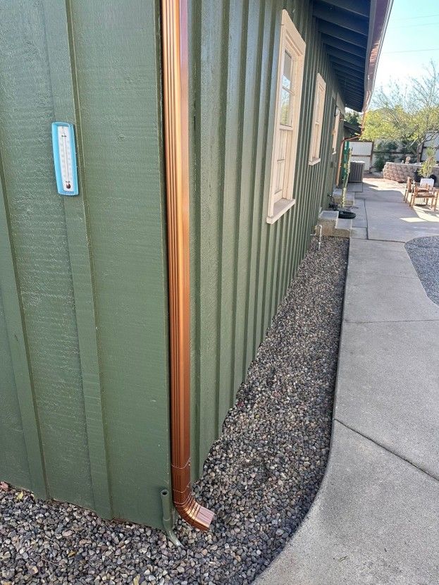 Green exterior wall with copper downspout beside a concrete walkway and gravel border
