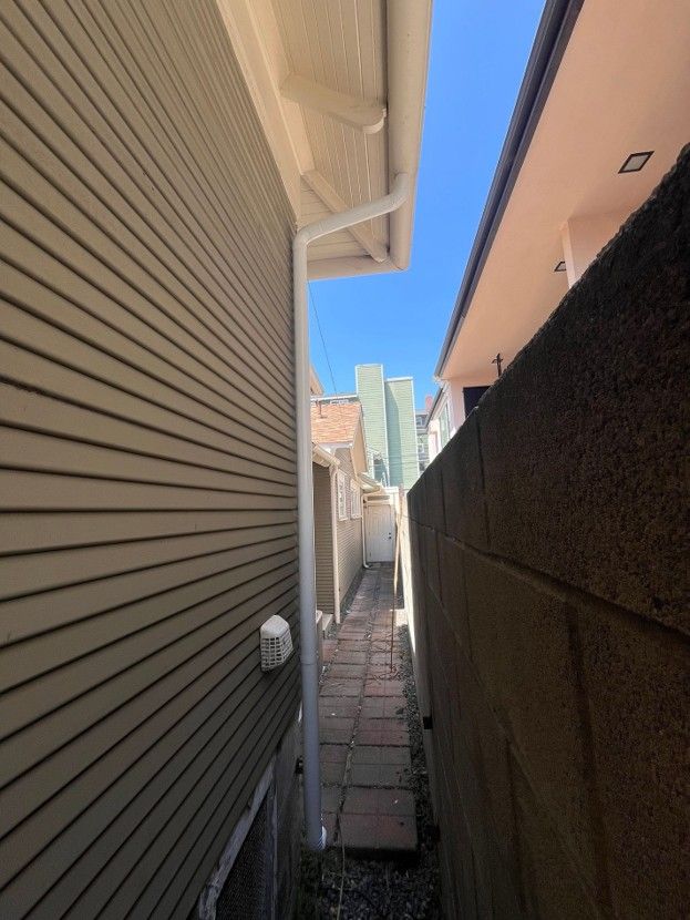 Narrow alley between a beige house and dark wall, with a clear blue sky and buildings in the distance.