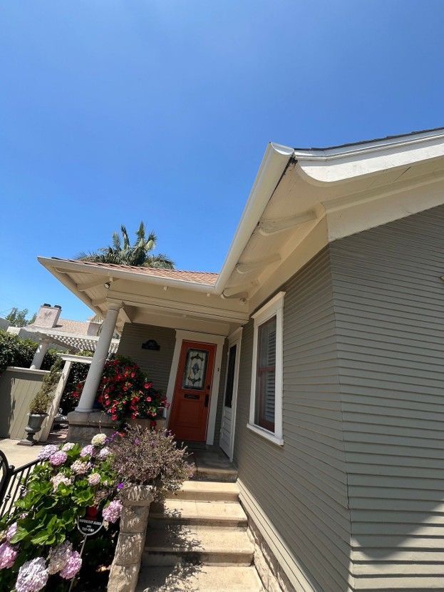 Front porch of a light gray house with steps, white columns, and colorful flowers under a blue sky