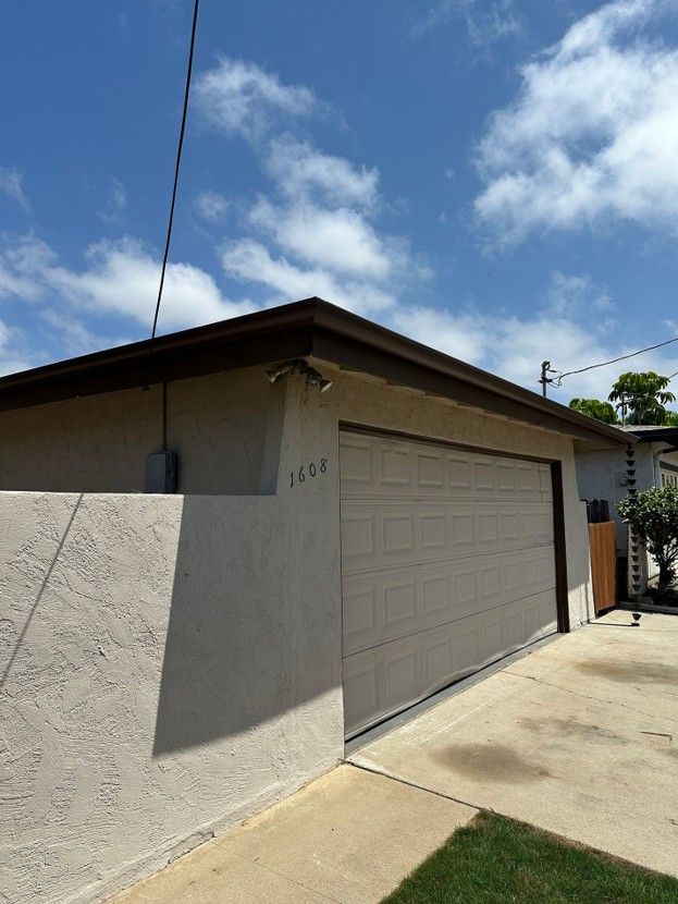 Single-story house exterior with beige garage door, stucco wall, and driveway under a blue sky