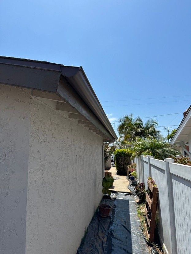 Narrow side yard between a stucco house and white fence, with potted plants and palm trees under a blue sky