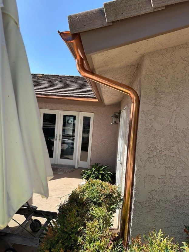 Copper downspout beside a stucco house courtyard with patio doors and potted plants