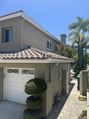 Two-story beige house with white garage, tiled roof, and trimmed topiary beside a narrow walkway