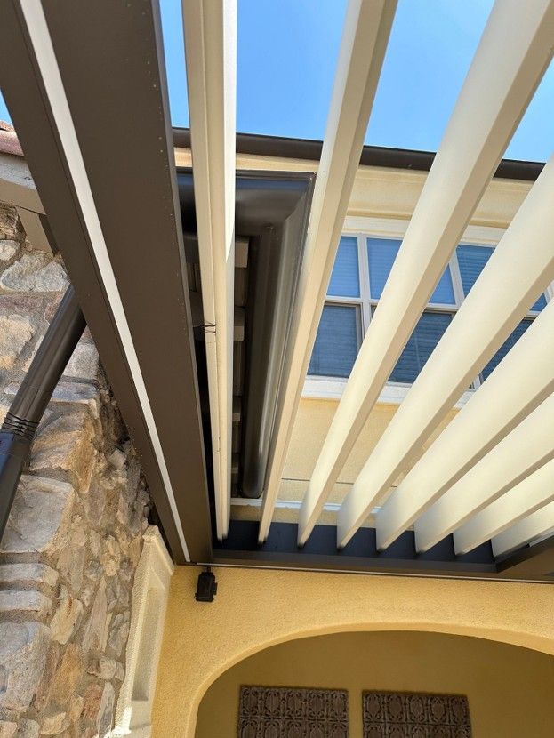 Looking up at a pergola with cream beams beside a stone wall and house eaves against a blue sky