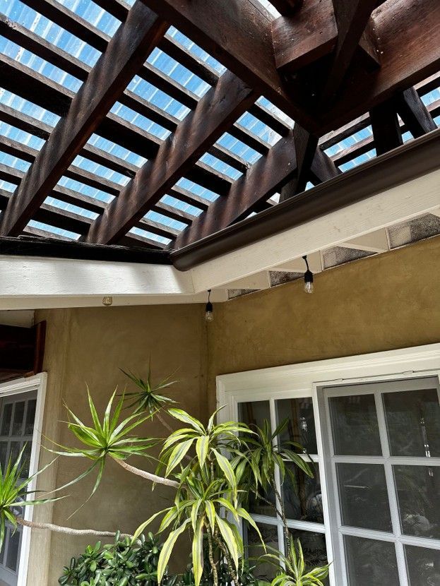 Patio with wooden pergola roof, white windows, beige wall, and potted plants in sunlight