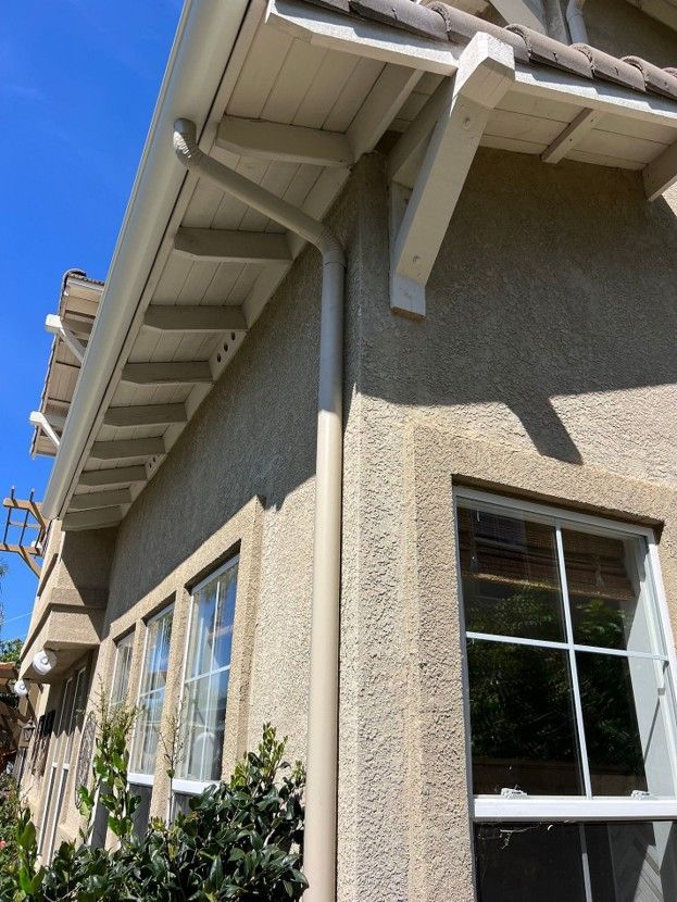 Exterior corner of a beige stucco house with white eaves and windows under a blue sky
