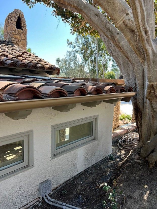 White stucco house with red tile roof beside a large tree in a sunny yard