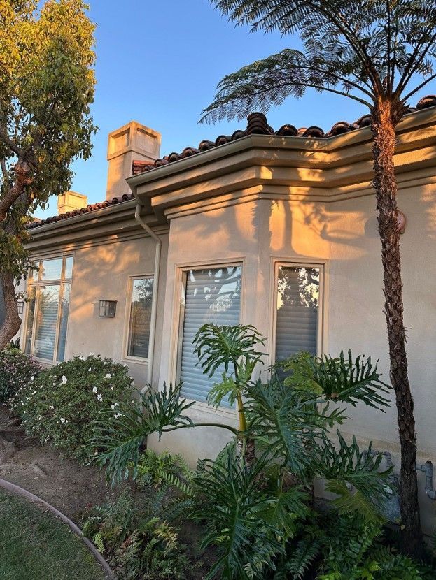 Single-story house with beige stucco, white shutters, tiled roof, and lush garden plants at sunset