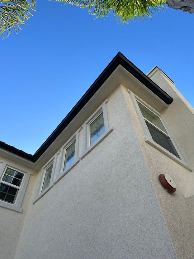 Low-angle view of a white house corner with black roof trim against a blue sky, framed by tree leaves