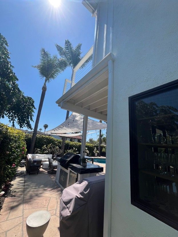 Sunny patio with palm trees, outdoor seating, and a covered grill beside a white house