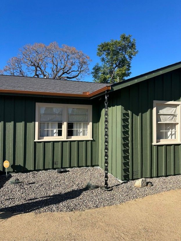 Green house exterior with white-trimmed windows and gravel yard under a clear blue sky