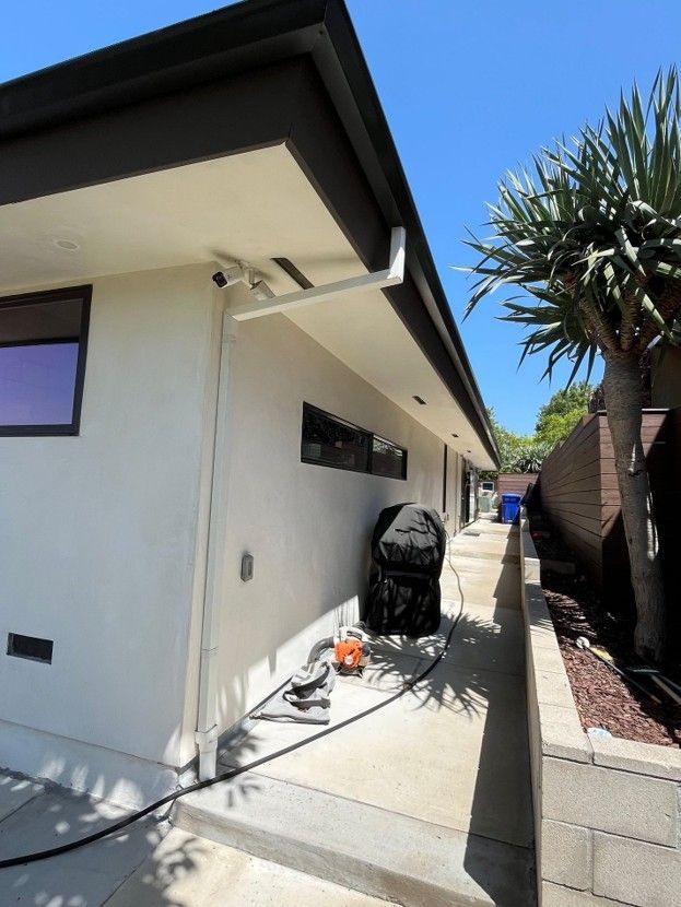 Side yard beside a white house with patio items, a grill, and a palm tree under a clear blue sky