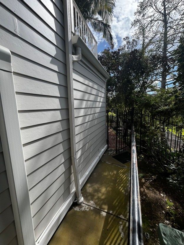 Narrow side yard beside a white house with siding, a metal railing, and trees beyond.