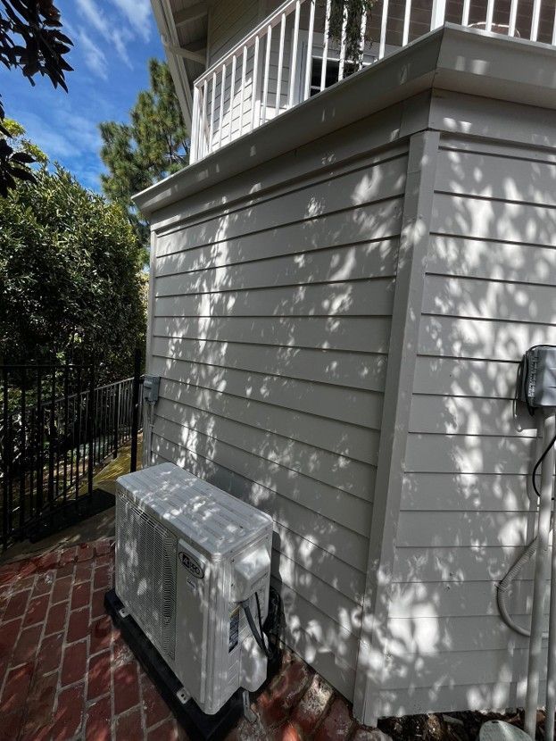 Side view of a light gray house exterior with a utility meter, black fence, and dappled tree shadows