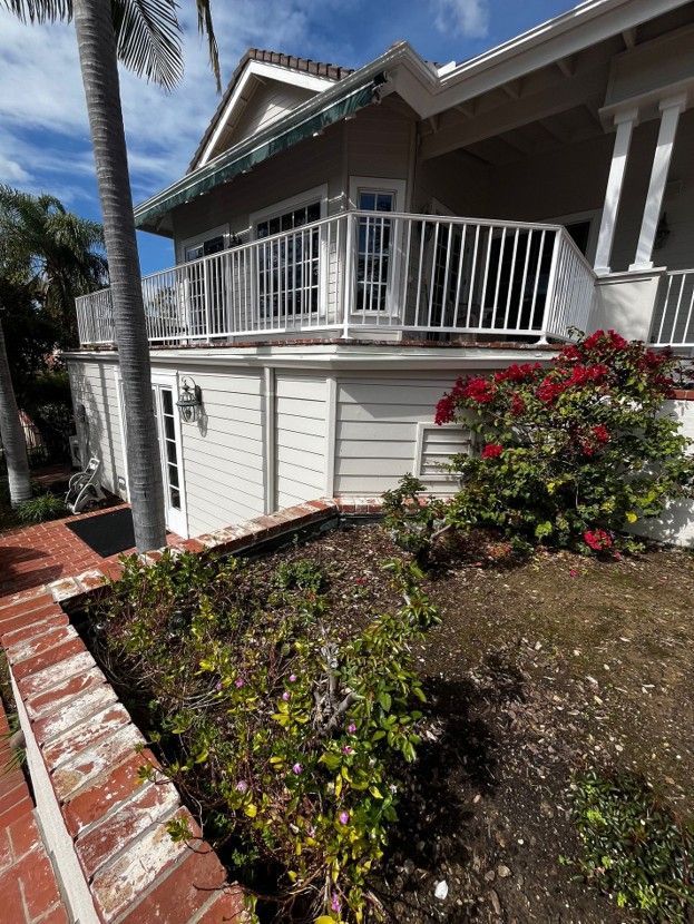 White porch with railing beside a red-brick walkway and flowering shrubs under a blue sky