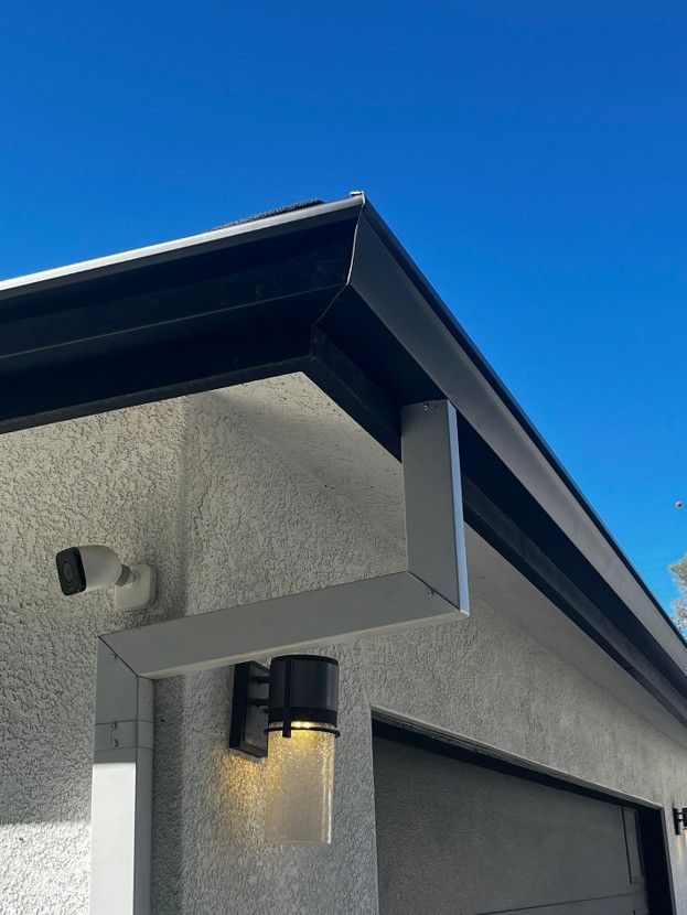 Gray stucco house corner with black roof trim and a lit outdoor wall lantern under a blue sky