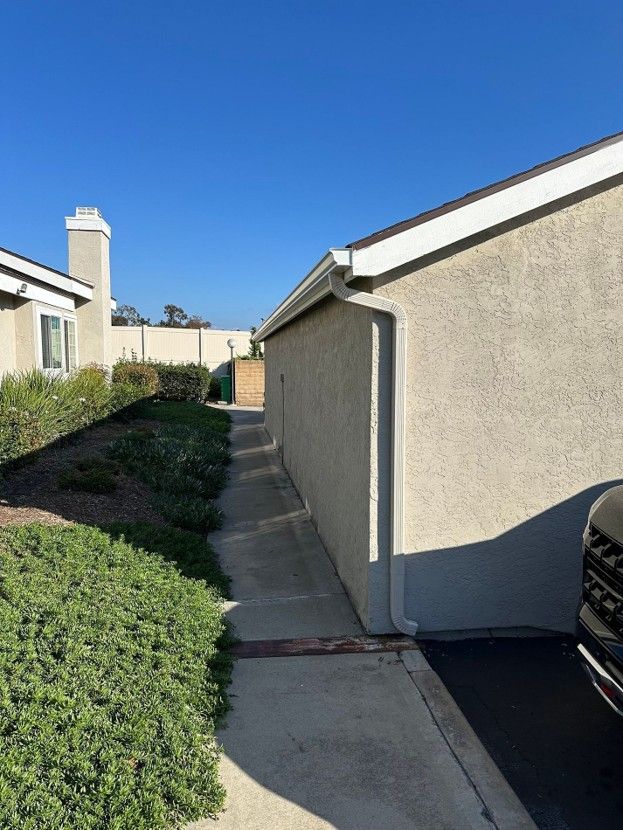 Narrow concrete side yard beside beige houses under a clear blue sky, with bushes and a parked car edge
