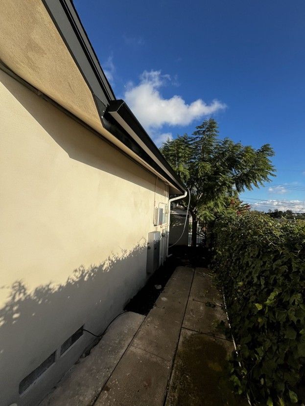 Narrow side yard beside a cream house, with a concrete path, tree, and clear blue sky.