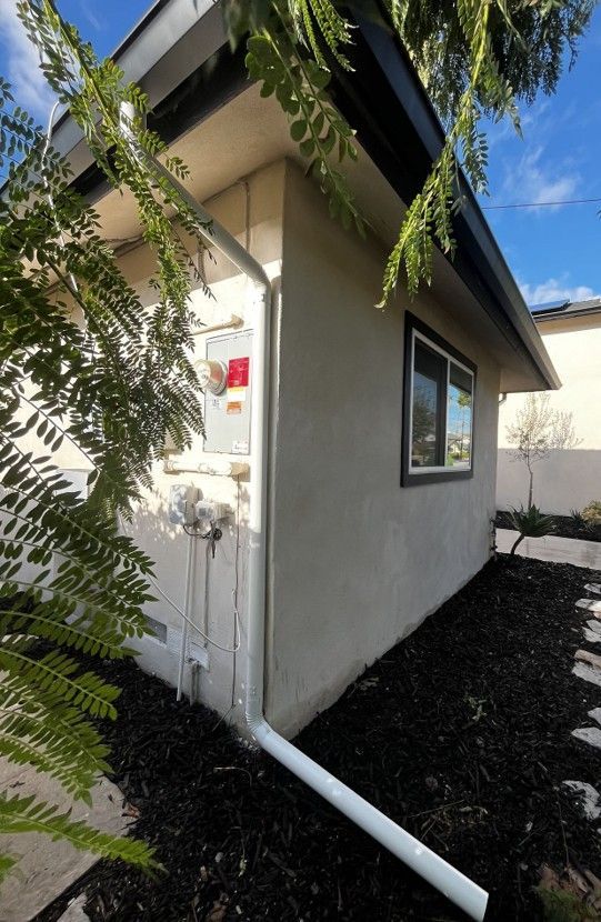 Side view of a small house exterior with white siding, black roof trim, and white drainage pipes.