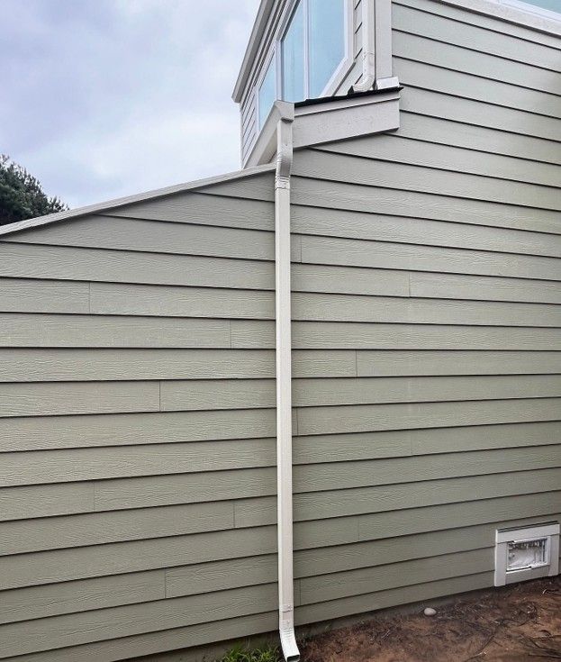 Side of a gray house with white trim and a downspout along the wall near a small basement vent.