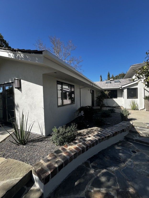 White stucco house exterior with black windows, desert landscaping, and a curved brick-edged walkway under a blue sky