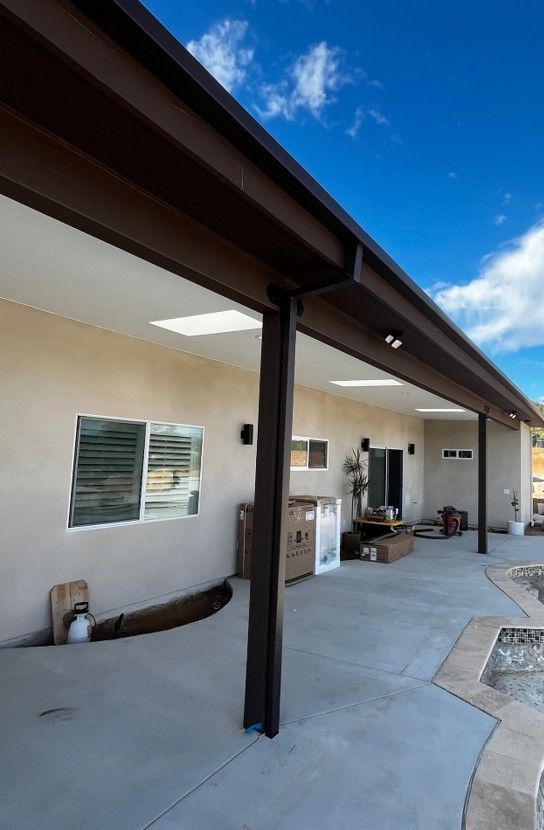 Covered patio beside a beige house with black support posts, windows, outdoor items, and blue sky.