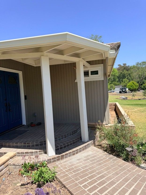 Front porch of a small building with white posts, brown siding, and brick steps in a sunny yard