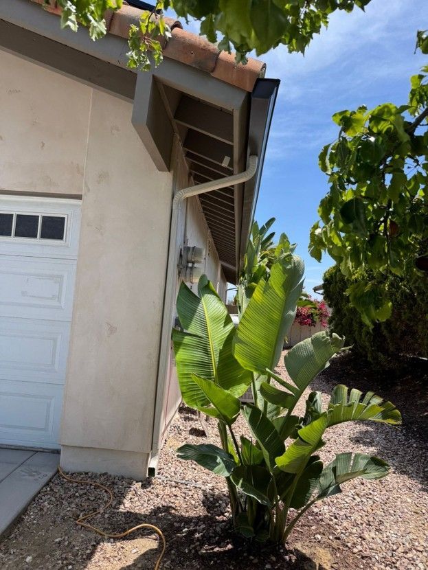 Side of a beige house with a garage, overhanging roof, and a banana plant in a gravel yard.