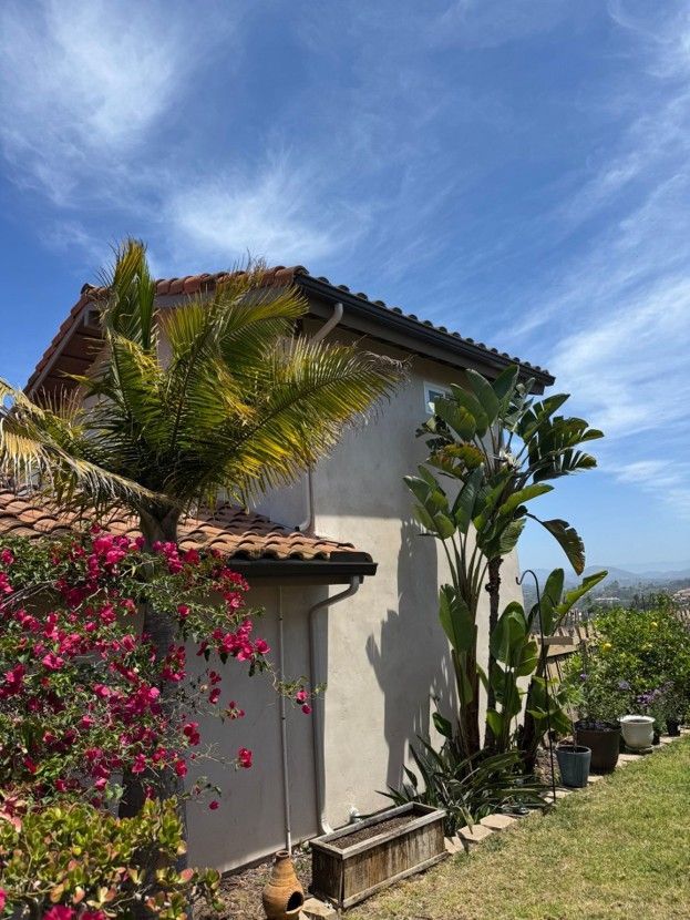 Two-story stucco house with a tiled roof, palm and flowering plants, under a bright blue sky