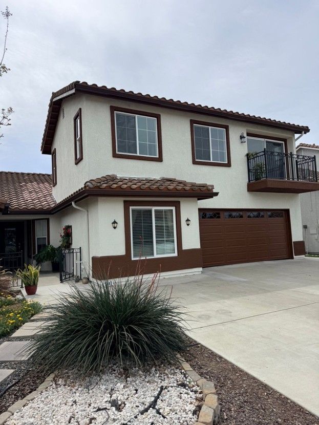 Two-story tan house with brown trim, front balcony, garage, and desert landscaping under a cloudy sky