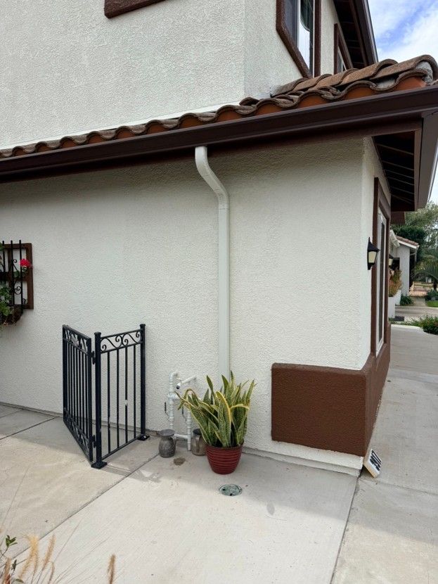 Side of a house with a tiled roof, white stucco wall, small black gate, and potted plant on a concrete patio.