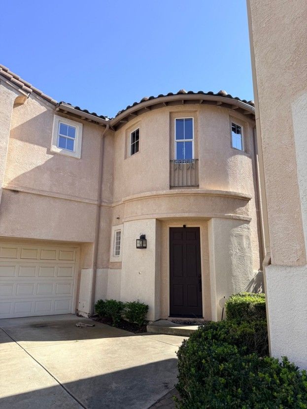 Two-story beige stucco townhouse with a garage and dark front door under a tiled roof.