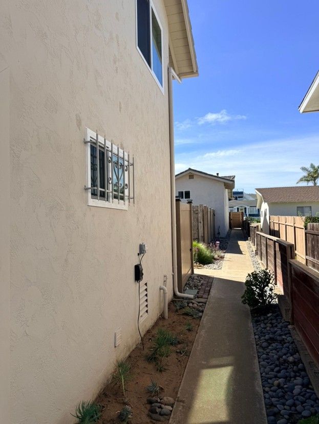 Narrow side yard walkway beside beige houses, with gravel, potted plants, and a fence under a blue sky