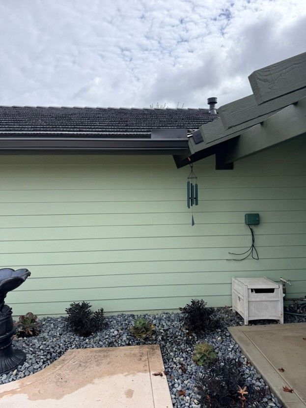 Exterior of a house with pale green siding, a black roof, and a small landscaped yard with rocks and shrubs