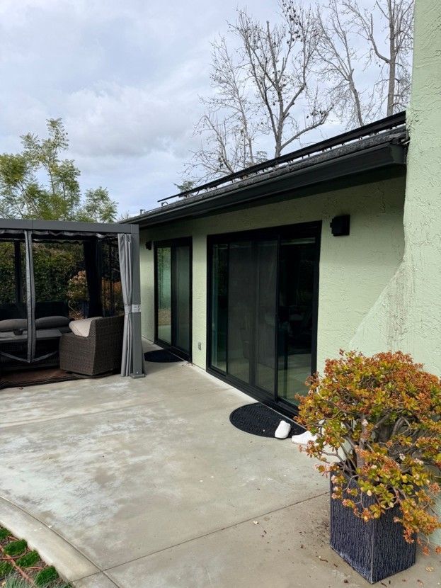 Patio with concrete floor, sliding glass doors, pergola seating, and potted plants beside a pale green house