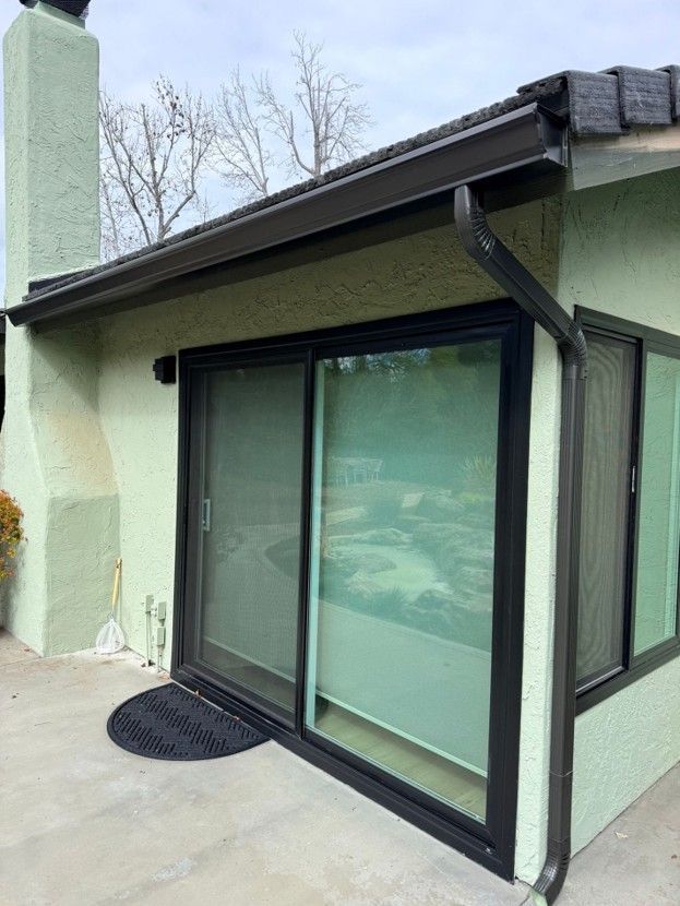 Black-framed sliding glass door on a pale green house exterior with a concrete patio and small doormat.