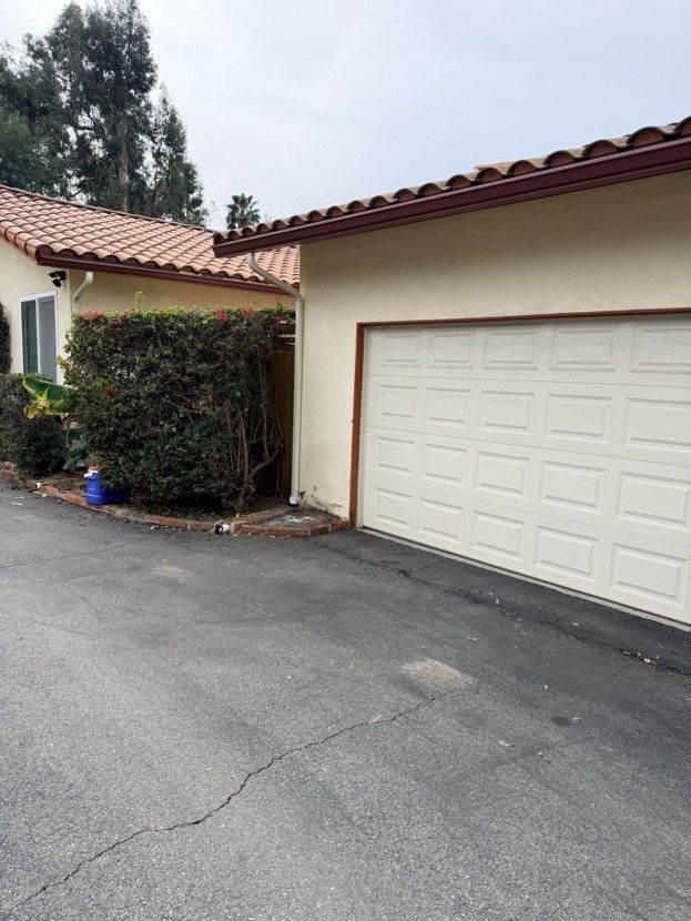 Driveway beside a beige garage with red tile roof and shrubs in front of a house