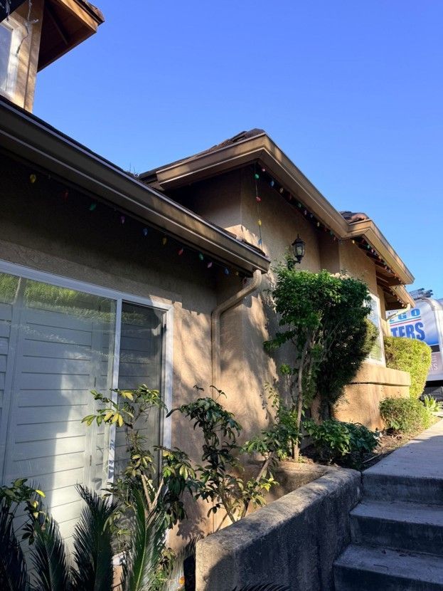 Stucco house exterior with white shutters, tiled roof, and a sunlit walkway lined with shrubs