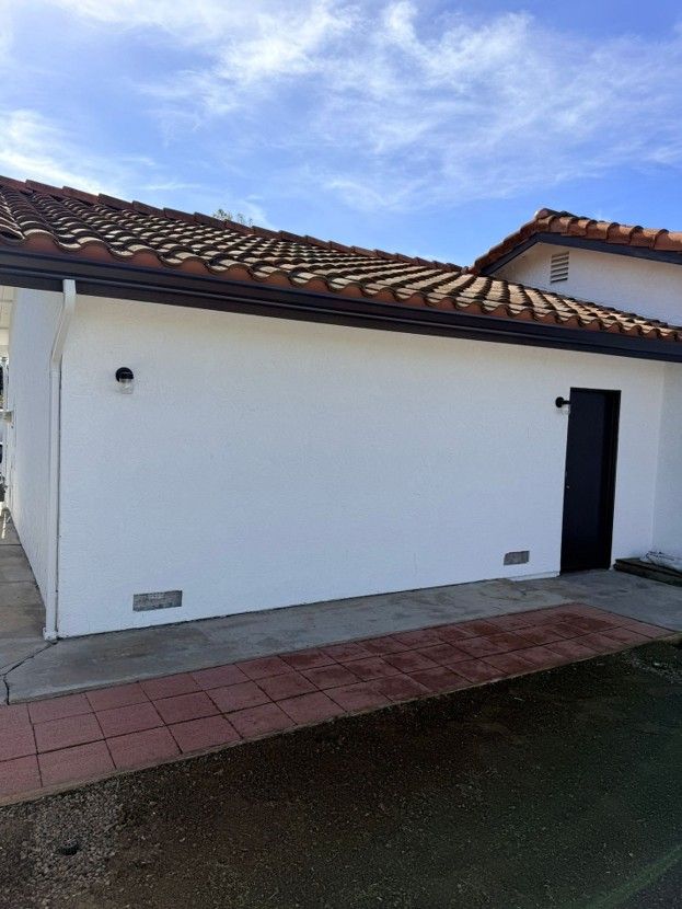 White stucco house exterior with red-tiled roof, dark doorway, and a paved walkway under a blue sky