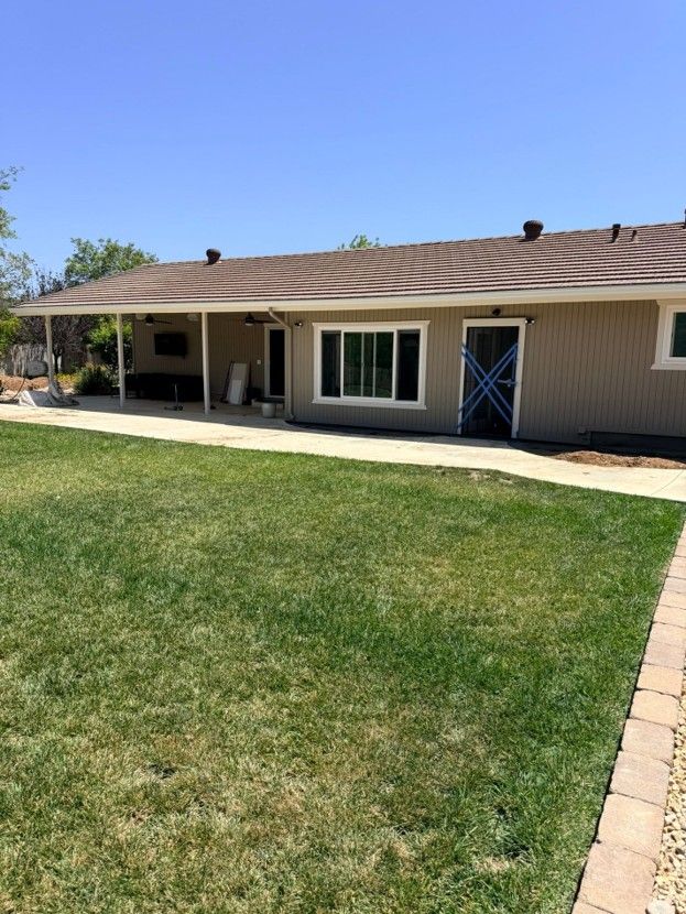 Single-story beige house with a green lawn, covered patio, and brick walkway under a clear blue sky
