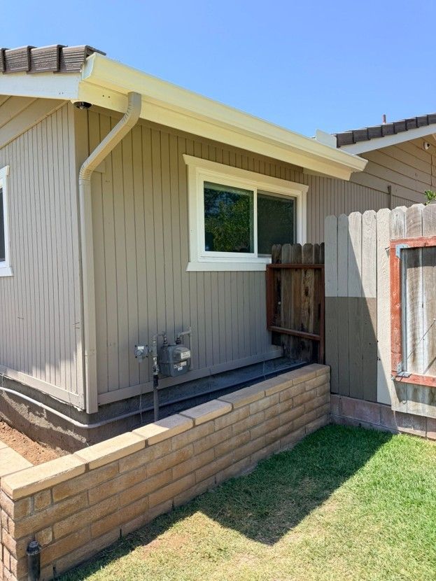 Side of a beige house with a small window, brick planter wall, fence, and patch of grass in a sunny yard