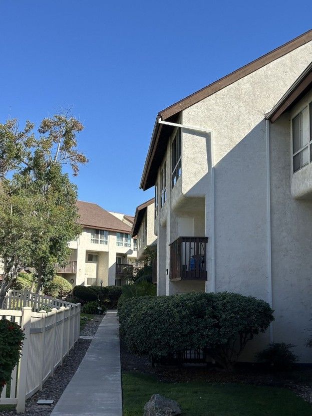 Apartment walkway beside beige buildings and hedges under a clear blue sky