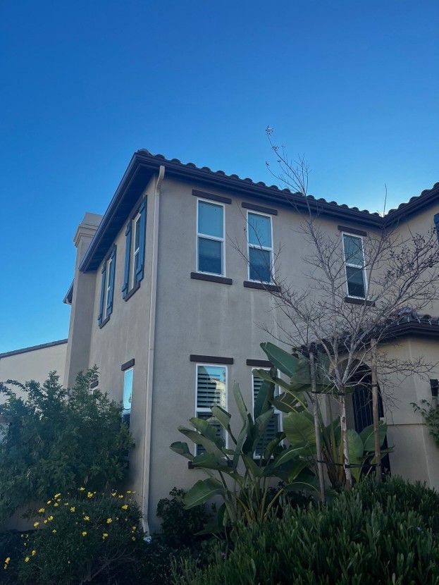 Two-story stucco house with blue shutters and a tiled roof, framed by lush greenery under a clear blue sky