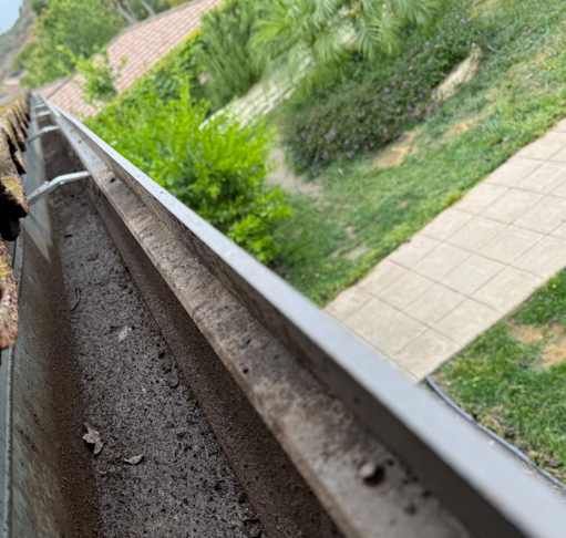 Close-up of a roof gutter with debris, overlooking a green hillside and tiled walkway below