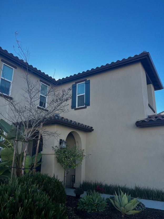 Stucco two-story house with blue shutters and a tiled roof, viewed from a garden under a clear blue sky